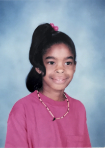 A young girl in a school photo. She's wearing a pink shirt and has a bow in her hair.