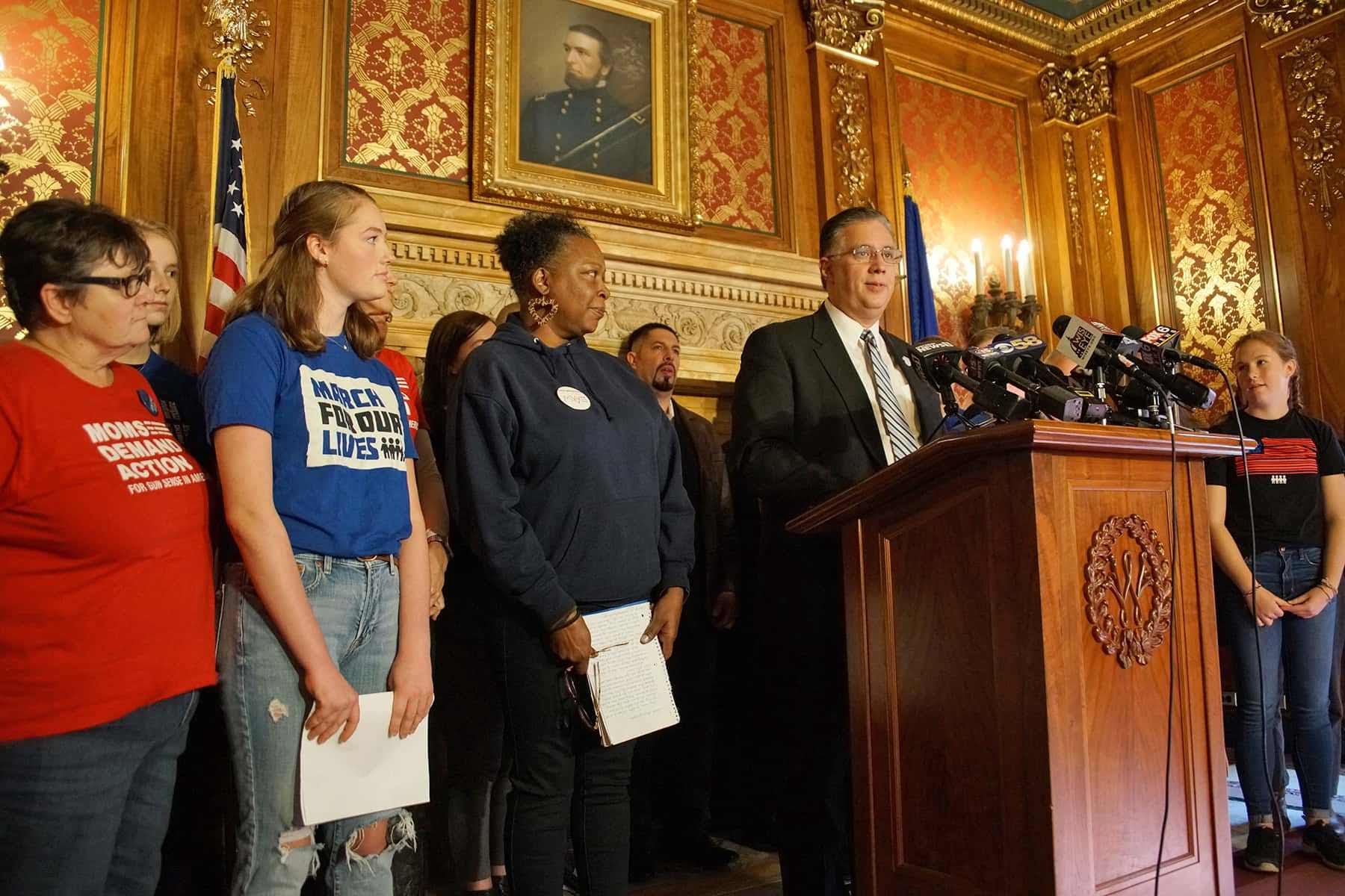 People lined up next to a podium with a man in a suit speaking at microphones, two on the edge are wearing red and black t-shirts with slogans on them. There is a historical painting behind them.