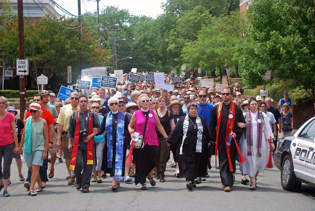 Religious leaders march in front of a crowd down a street, with trees behind them and a police vehicle in the corner of the image.