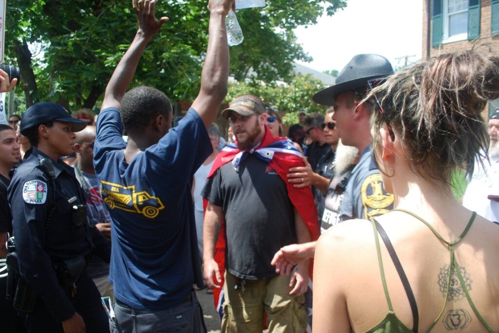 A man in a blue T-shirt and raised arms faces another man who has a confederate flag draped around his shoulders. On the side, an officer in a baseball cap is looking at the two men, and another officer is on the other side. A woman’s back is in the frame as well.