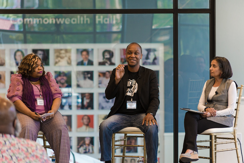 Three people in chairs on a stage, man in middle speaking with microphone