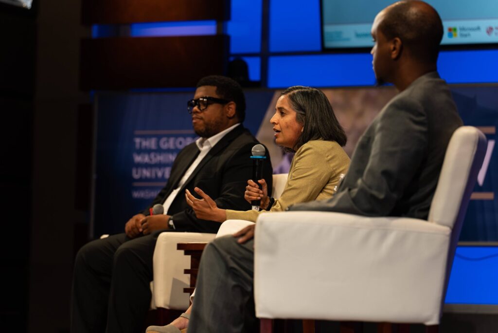 Three people on a stage, seated in white armchairs. Person in the middle speaks into a microphone.