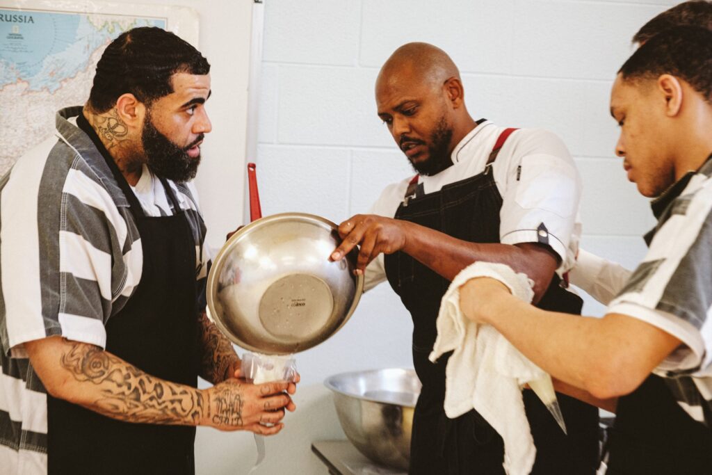 At the center of the photo a man in a white chef's coat stirs something in a stainless steel bowl. On either side of him, two men in black aprons over black and white striped jumpsuits watch what he is doing.