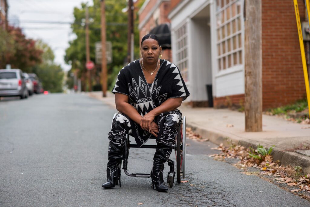 Woman in black and white and boots looks at the camera, in a wheelchair on a street, with buildings and cars in the background.