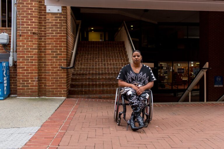 A woman in a wheelchair is photographed on a brick sidewalk with a stairwell behind her.