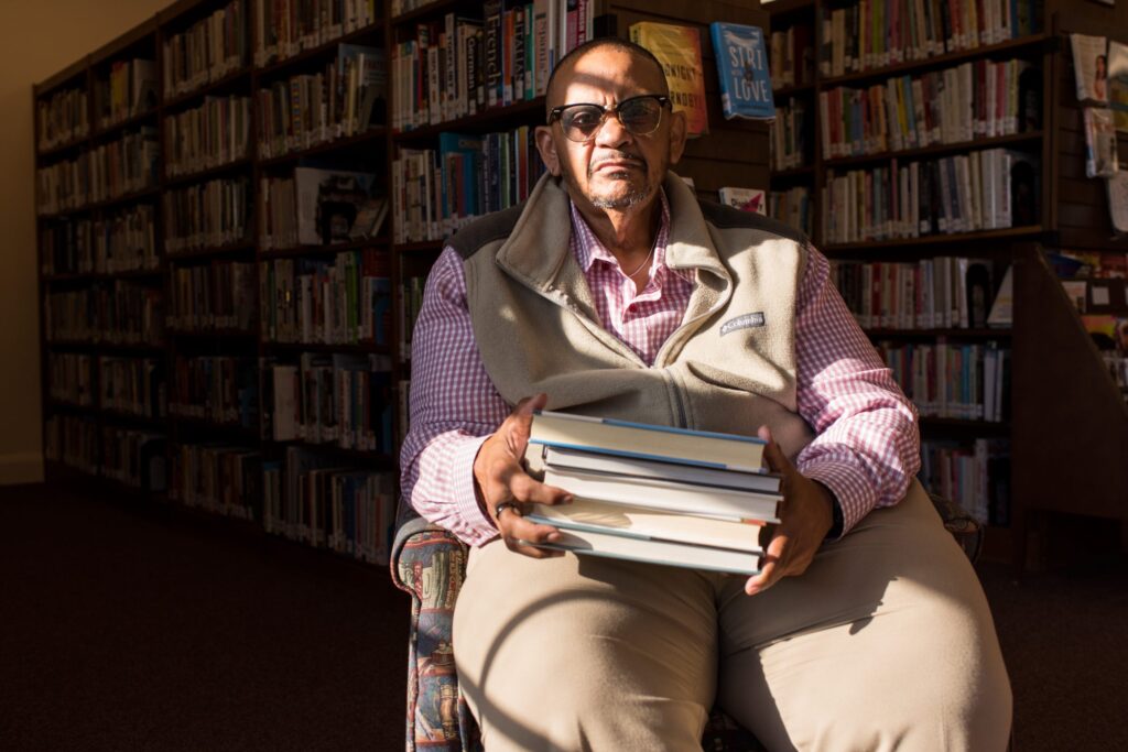 A person in a chair holding a stack of books, in front of shelves of books, with a band of light across his face.