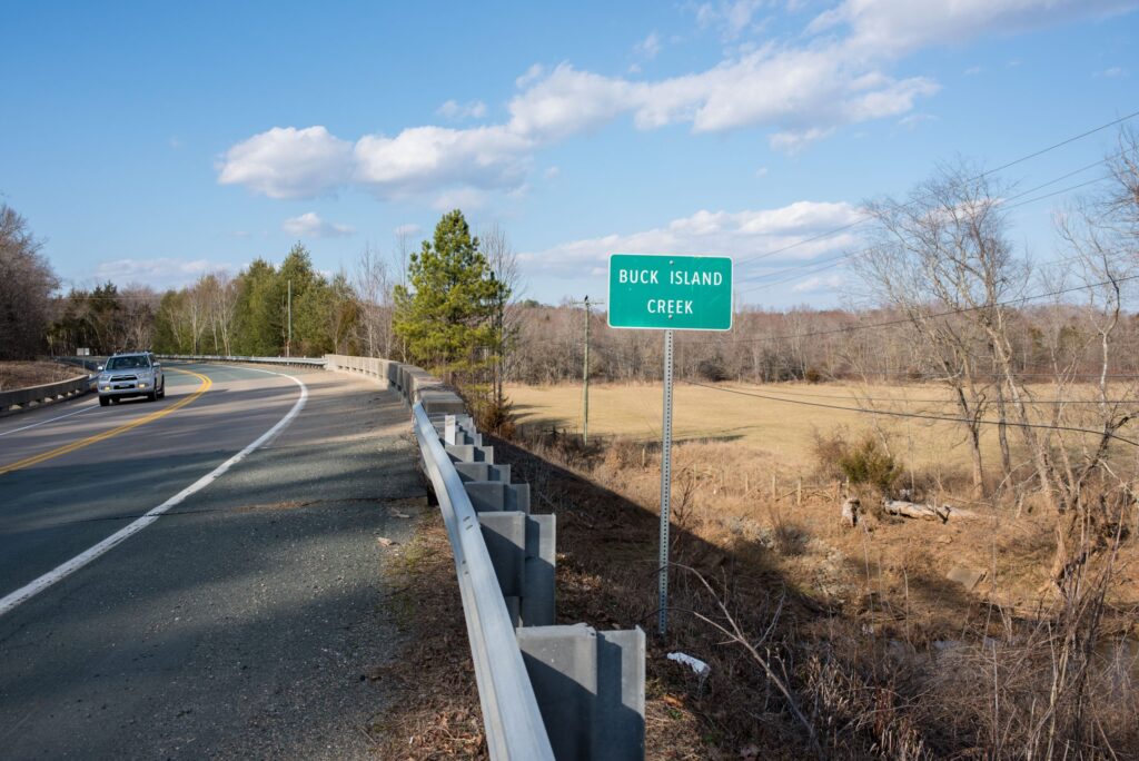 A two-lane road with a car approaching, cleared land and some trees to the side, white clouds in a blue sky. In the center of the image, a green road sign says, "BUCK ISLAND CREEK."