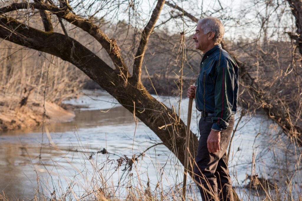 A man with a walking stick looks out over a river, trees behind him.