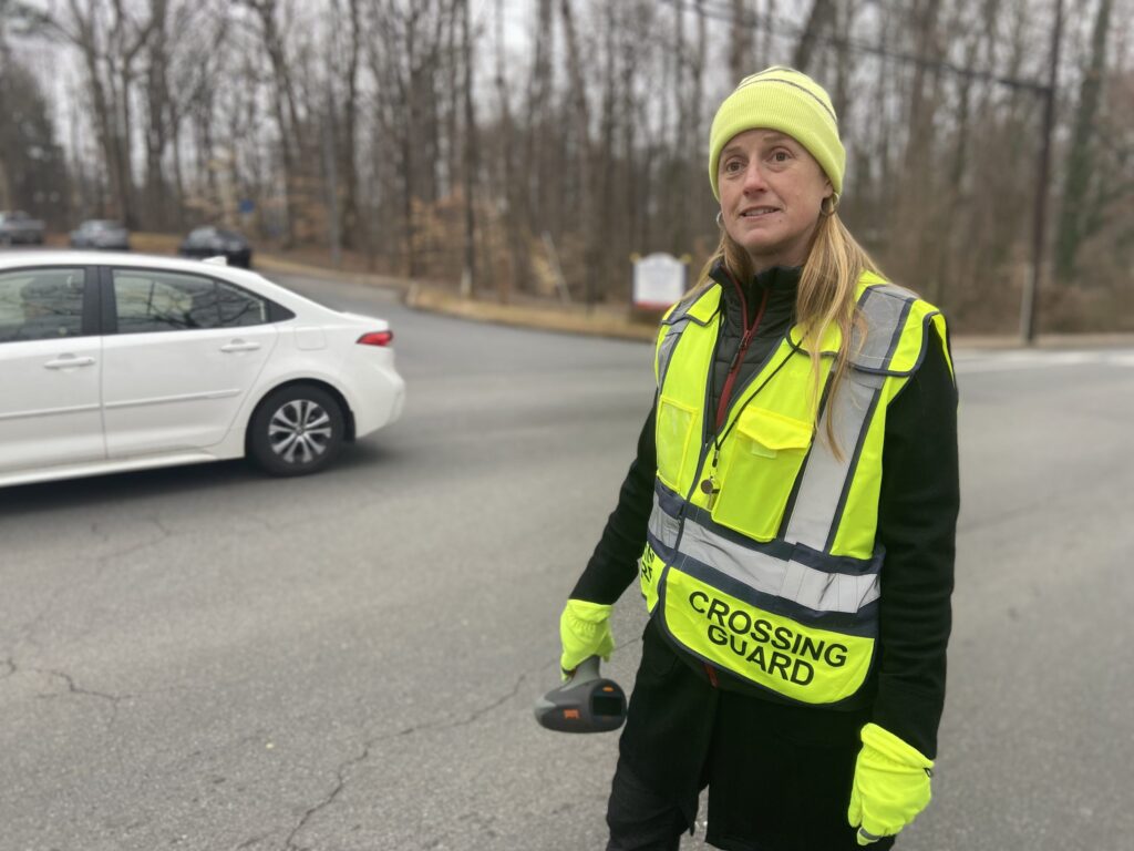 Person with long hair and beanie, bright yellow vest that reads "CROSSING GUARD" stands in an intersection with a car passing, radar gun in hand.