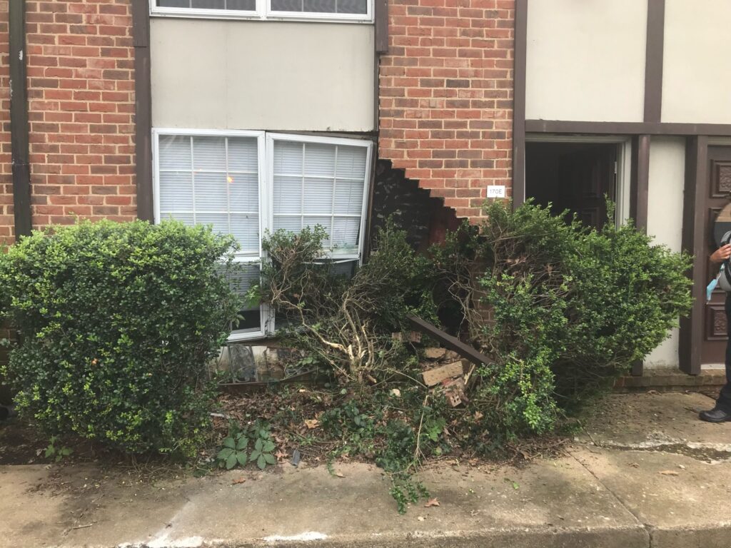 A brick and cement building has a large hole in the front between a door and windows, with a broken bush covering part of the door, a person in partial view to the side.