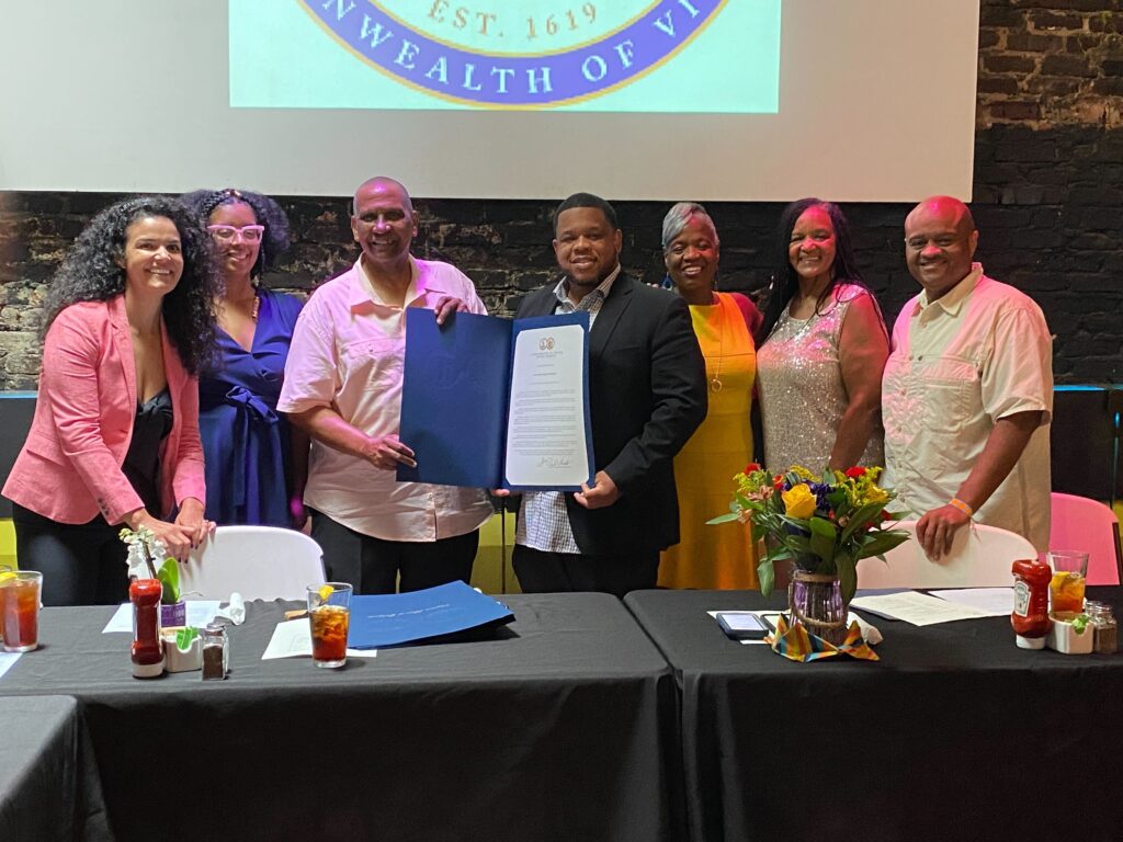 Seven people behind a table, with flowers in front. Two in the middle are holding open a certificate. A logo is protected behind them, the bottom of the seal of the Commonwealth of Virginia.