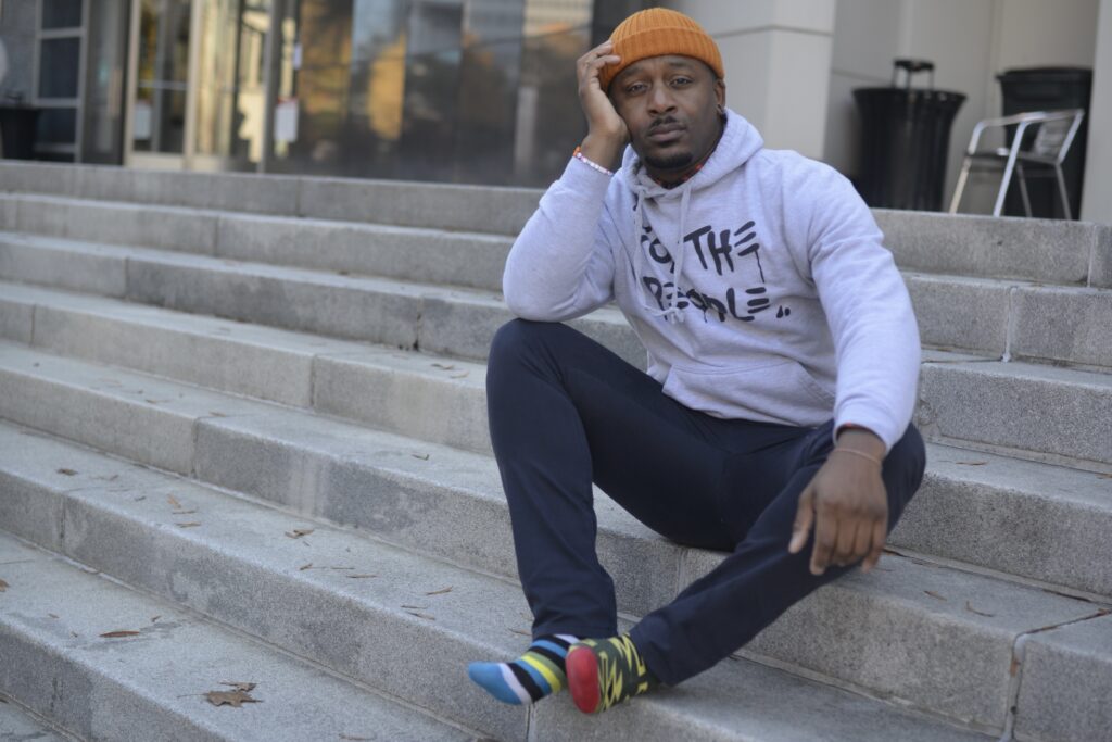 Man in hoodie and beanie sits on outdoor stairs with his head leaning against his hand.