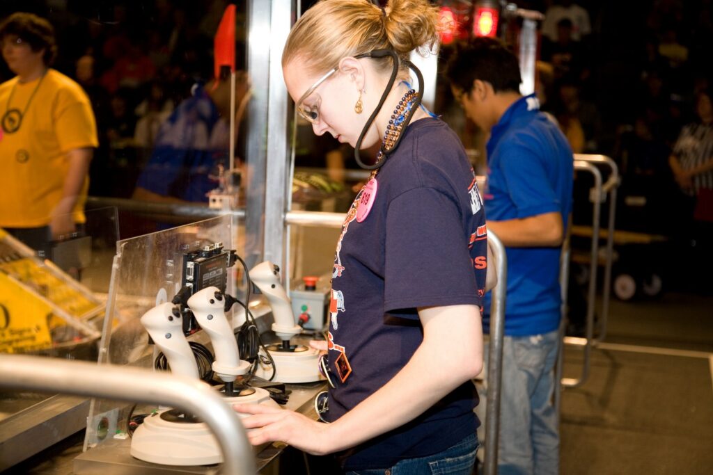 A young girl wearing safety glasses stands at a row of controllers, wires and tech. Two other young people are behind her, and they appear to be in front of an audience.